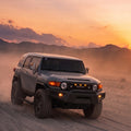 A dark gray off-road SUV driving on a dusty desert road at sunset, with mountains in the background and the vehicle's headlights and fog lights turned on.